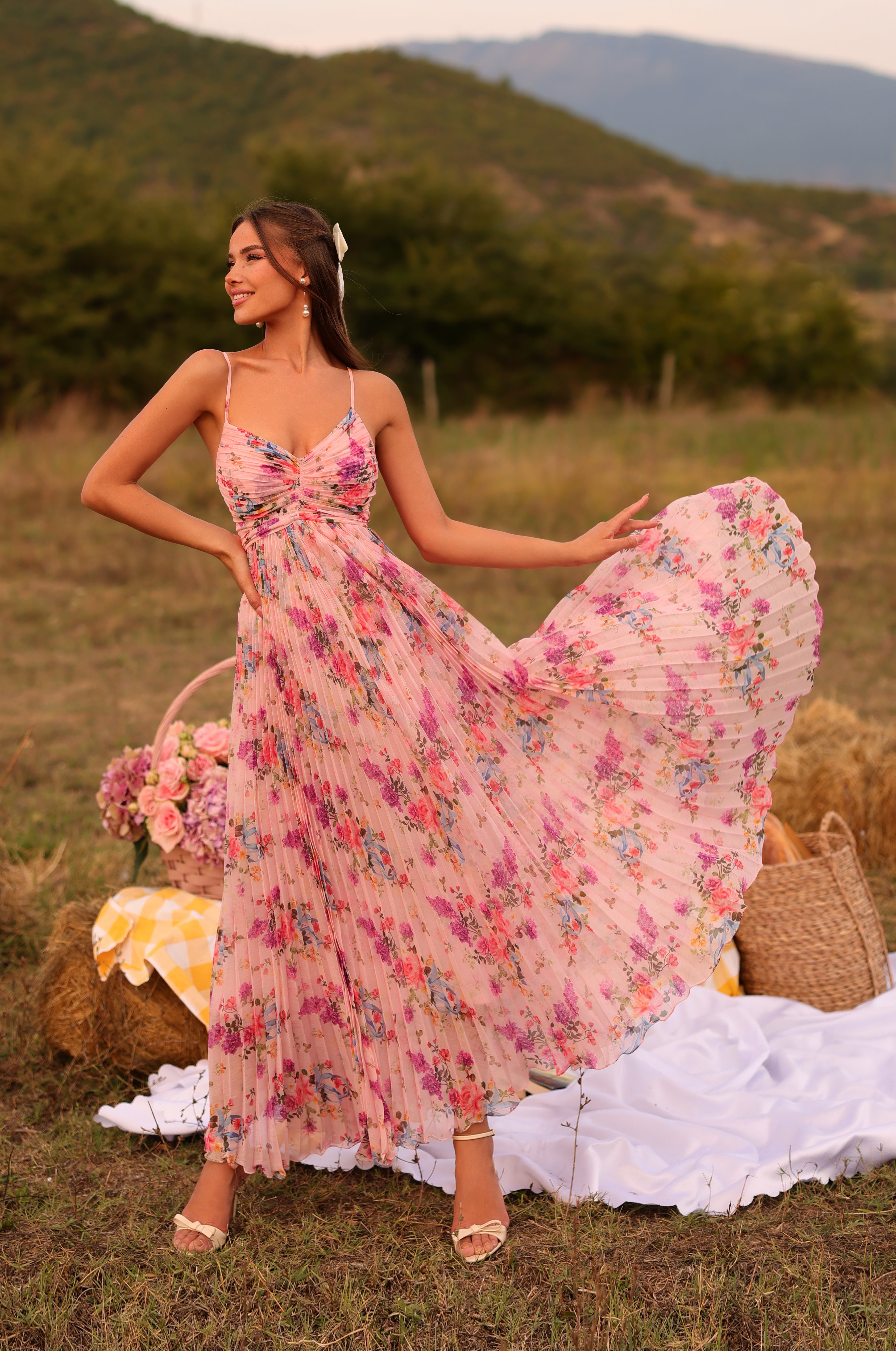 A woman poses outdoors on grass with hills in the background, wearing the Pleated Blush Pink Floral Maxi Dress: Florinne. Nearby, picnic items, flowers, and a blanket complete the scenic setup.